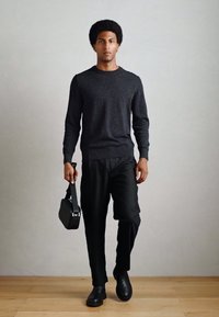 Black sweater, black trousers, and black shoes. Model holds a small black bag, standing on a wooden floor with a neutral background.