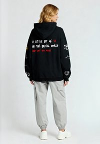 Juoda gobtuvinė džemperis su grafiniu tekstu nugarinėje pusėje. Turi perteklinį kirpimą, įsegamus rankovėmis ir derinamas su šviesiai pilkomis krovinių kelnėmis bei sportbačiais.