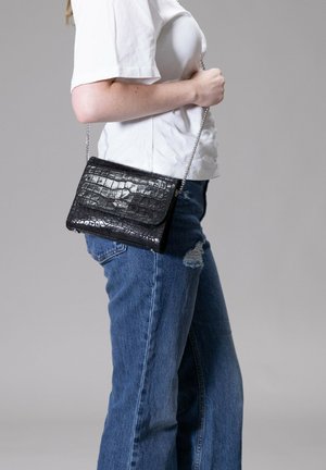 Schwarze Kunstleder-Crossbody-Tasche im Krokodil-Look mit glatter Textur, rechteckiger Form und silberner Kettenriemen, getragen zu lässigen blauen Jeans.