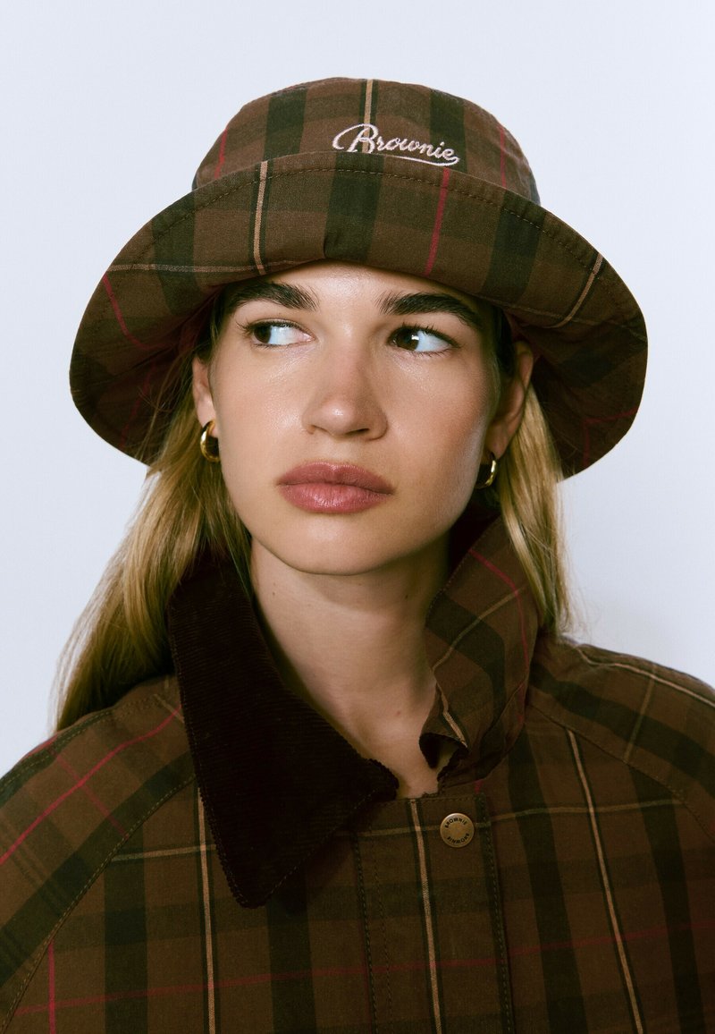 Joven mujer que lleva un sombrero de cubo a cuadros marrón y una chaqueta a juego con cuello alto, mirando de lado, con pendientes de aro dorados visibles.