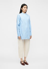 Γυναίκα με μακριά σκούρα μαλλιά φοράει μπλε-λευκό ριγέ μπλουζάκι με μακριά μανίκια, κρεμ παντελόνι με φαρδιά πόδια και καφέ σκούρα παπούτσια Mary Jane, όρθια.