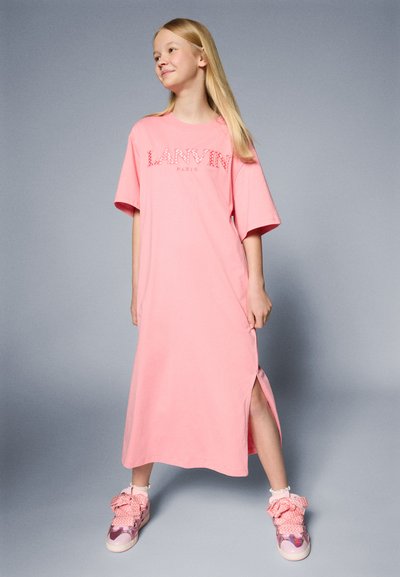 Vestido de camiseta de algodón rosa oversized con estampado de logo, con aberturas laterales, combinado con zapatillas rosas que tienen lazos decorativos y un acabado brillante.