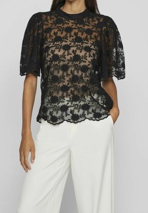 Femme portant un chemisier en dentelle florale noire transparente avec manches bouffantes et col montant, associé à un pantalon blanc à jambes larges, debout la main dans la poche.