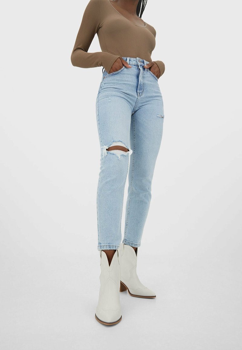 Ljusblå högmidjade jeans med slitna knäpartier, parat med vita cowboystövlar med spetsiga tår och dekorativ sömnad.