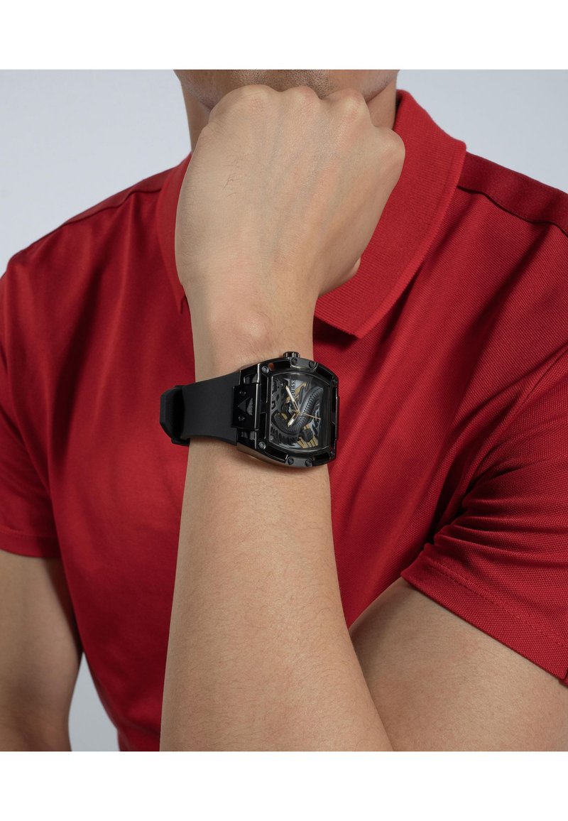 Black rectangular watch with a patterned face, black silicone strap, and metal accents, worn on a wrist. Man's red polo shirt in background.