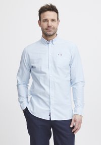 Lys blå, langermet skjorte med knapper, hvite striper, brystlomme og liten logo. Materialet ser glatt og uformelt ut.