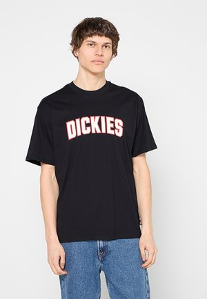 Joven con cabello corto y castaño que lleva una camiseta negra Dickies y vaqueros azules, de pie frente a un fondo blanco.