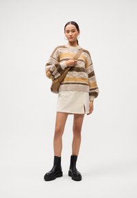 Femeie îmbrăcată cu un pulover striped maro și beige, fustă mini crem cu fanta, botine negre cu talpă groasă, cercei rotunzi și o poșetă maro purtată pe umăr.