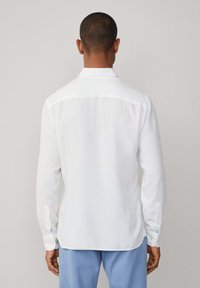 Man met kort haar, gekleed in een witte blouse met lange mouwen en lichtblauwe broek, staand met de rug naar de camera tegen een effen achtergrond.