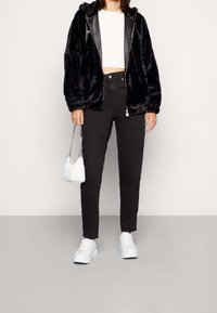 Mujer vistiendo una chaqueta de piel sintética negra, un top corto blanco, jeans negros de tiro alto, zapatillas blancas y sosteniendo un pequeño bolso blanco con correa de cadena.