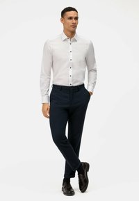 Weißes Langarmhemd mit schwarzen Knöpfen, kombiniert mit dunkelblauen Hosen. Modell trägt schwarze Schuhe und präsentiert einen sauberen, maßgeschneiderten Look.