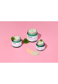 Trzy słoiki kremu Dr. Jart+ Cicapair w różnych rozmiarach, z zielonym i białym opakowaniem. Obok nich znajduje się jasnozielony próbka produktu.