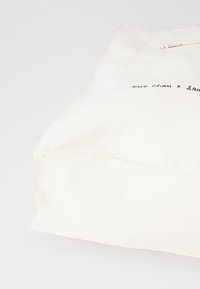 Cream-colored canvas bag with a flat shape, featuring a front pocket and zipper closure. Signed with "Kris Chan x" in black.