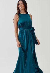 Teal satin maxi dress with a round neckline, side tie accent, and flowing skirt featuring draped texture. Smooth, lightweight fabric.