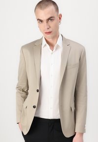 Bežinis švarkas su siauru pjūviu, vienu mygtuku ir krūtine kišene. Užrišamas ant baltos marškinių, derinamas su juodomis kelnėmis. Lygus paviršius.