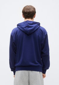 Tengerészkék kapucnis pulóver, puha anyagból készült, laza szabással és bordázott mandzsettával, hátulnézetben. Alatta szürke melegítőnadrág található.