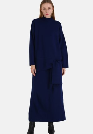 Navy blue knitted dress with a high neck and long sleeves; features a tie detail at the waist and a relaxed fit, extending to the ankles.