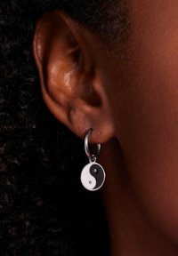 Yin-yang oorbellen met een zilveren hoop, voorzien van een zwart-witte ronde hanger met twee kleine edelstenen. Krullend haar zichtbaar op de achtergrond.