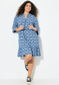 Donna con capelli ricci che indossa un vestito blu a fantasia e sneakers blu scuro, tiene degli occhiali da sole e si appoggia a un muro bianco.