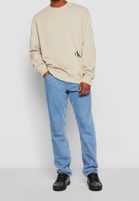 Sweatshirt en coton beige avec col ras du cou et logo noir brodé sur le côté, associé à un jean en denim bleu clair et des baskets noires.