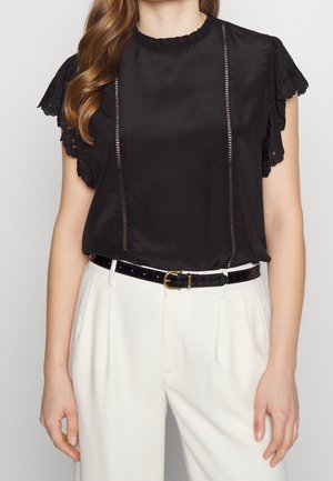 Femme portant un chemisier noir à manches en dentelle rentré dans un pantalon blanc taille haute avec une ceinture noire à boucle dorée.