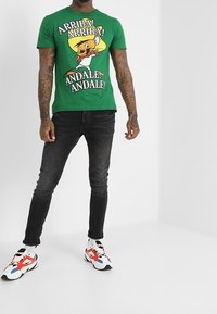 Zielona bawełniana koszulka z nadrukiem postaci z kreskówki w żółtym i czarnym tekście. Połączona z czarnymi obcisłymi dżinsami i wielokolorowymi trampkami.