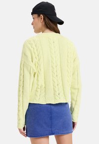 Frau trägt einen pastellgelben Strickpullover, einen blauen Denim-Minirock und eine schwarze rückwärts gerichtete Cap, mit dem Rücken zu einem weißen Hintergrund.