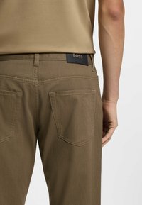 Primo piano di una persona che indossa pantaloni marroni e una camicia beige, mostrando le tasche posteriori e un'etichetta nera con la scritta "BOSS" sulla fascia della vita.