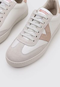 Zapatillas blancas con acentos de ante gris claro, que presentan una punta redondeada, un diseño de cordones y un logo en la lengüeta. Detalles de superficie texturizada visibles.