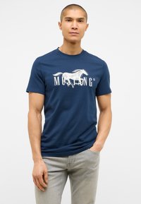 Námořnická modrá bavlněná tričko s grafickým motivem bílého koně a textem "MUSTANG". Krátké rukávy a kulatý výstřih. V kombinaci se světle šedými džíny.