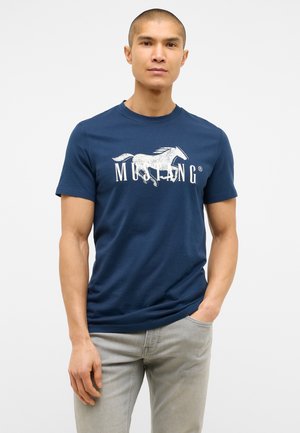 Námořnická modrá bavlněná tričko s grafickým motivem bílého koně a textem "MUSTANG". Krátké rukávy a kulatý výstřih. V kombinaci se světle šedými džíny.