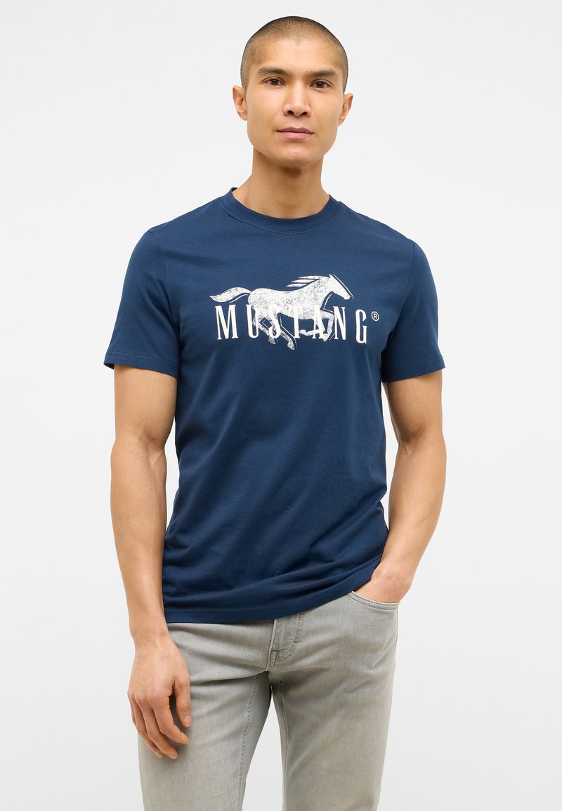 Námořnická modrá bavlněná tričko s grafickým motivem bílého koně a textem "MUSTANG". Krátké rukávy a kulatý výstřih. V kombinaci se světle šedými džíny.