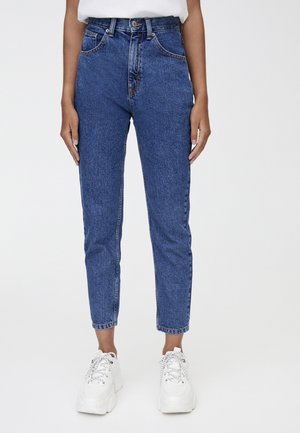 Person trägt hoch taillierte blaue Jeans und weiße klobige Sneakers, steht vor einem schlichten hellen Hintergrund.