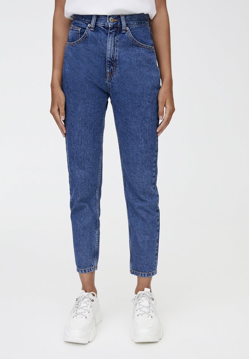 Personne portant un jean en denim bleu taille haute et des baskets blanches épaisses, debout devant un fond clair uni.