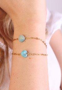 Gouden kettingarmband met twee hartvormige stenen: één lichtblauw en één turquoise, elk met unieke texturen en patronen.