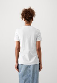 Person med lockigt hår i en vit t-shirt och ljusa jeanskjol står med ryggen mot kameran mot en enkel ljus bakgrund.