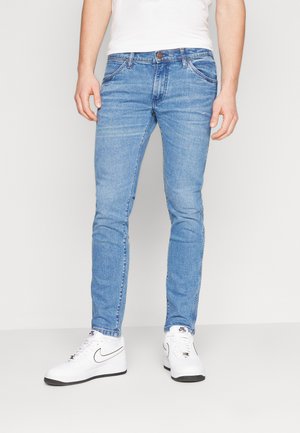 Jeans in denim blu chiaro con un design aderente, cuciture dettagliate e cinque tasche, abbinati a sneakers bianche per un look casual.
