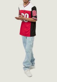 Camisa deportiva en rojo, negro y blanco con un gran '89' en el frente, combinada con jeans claros desgastados y zapatillas blancas.