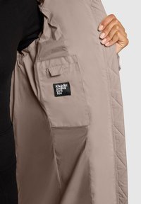 Gequiltede beige jasstof met een gladde textuur; heeft een grote binnenzak en een merketiket dat aan de binnenkant is gestikt.