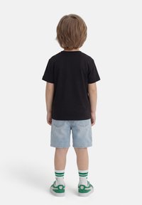 Παιδί με ανοιχτό καστανό μαλλί στέκεται με το πρόσωπο προς τα πίσω, φορώντας μαύρο μπλουζάκι, ανοιχτό μπλε τζιν σορτσάκι, λευκές κάλτσες με πράσινες ρίγες και πράσινα αθλητικά παπούτσια.