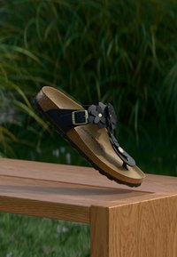 Μαύρο σανδάλι με διακοσμητικά λουλουδένια στοιχεία, ρυθμιζόμενο λουράκι, πάτο από φελλό και λαστιχένια σόλα, τοποθετημένο σε ξύλινη επιφάνεια.