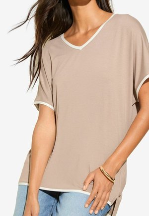 Beżowa koszulka z krótkim rękawem i dekoltem w serek, z białym wykończeniem na rękawach, dekolcie i dole, noszona z niebieskimi dżinsami i złotą bransoletką na lewym nadgarstku.