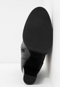 Bottine en cuir noir avec semelle texturée et petit talon, posée sur la pointe contre un coin blanc en arrière-plan.