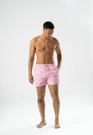 Mężczyzna bez koszulki, stojący boso, ubrany w różowo-białe pasiastye kąpielówki, z ręką w kieszeni, patrzący w dół na jednolitym białym tle.