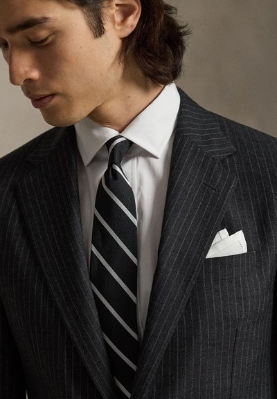 Άνδρας με σκούρο κοστούμι με κάθετες ρίγες και λευκό πουκάμισο φορά γραβάτα με διαγώνιες μαύρες και γκρι ρίγες και λευκό μαντηλάκι στην τσέπη, κοιτάζοντας προς τα κάτω αριστερά.