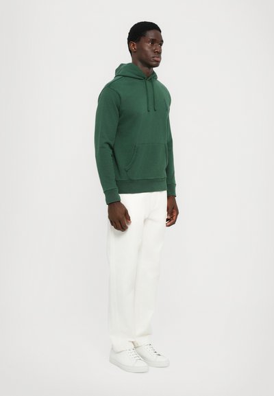 Grön hoodie med framficka och justerbara dragsnoddar, bärs med vita vida byxor och vita sneakers. Slät textur, avslappnad design.