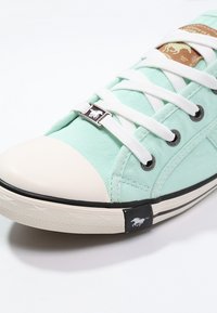 Hellgrüner Leinensneaker mit weißer Gummikappe vorne, schwarzer Sohle, silbernen Akzenten an den Schnürsenkeln und einem Logo an der Ferse.