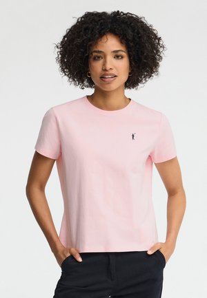 Kobieta z kręconymi włosami, ubrana w jasnoróżową koszulkę z krótkim rękawem, z małym ciemnym logo na piersi, ręce w kieszeniach ciemnych spodni.