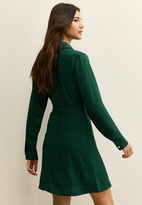 Zielona, długa sukienka z rękawami, zapinana w pasie, z kołnierzykiem i teksturowanym materiałem. Posiada mankiety z guzikami i sięga powyżej kolan.