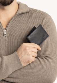 Schwarze Leder-Geldbörse, die in einer Hand gehalten wird, mit einem eleganten Design und strukturierter Oberfläche. Die Hand befindet sich gegen einen hellbeigen, gestrickten Pullover.